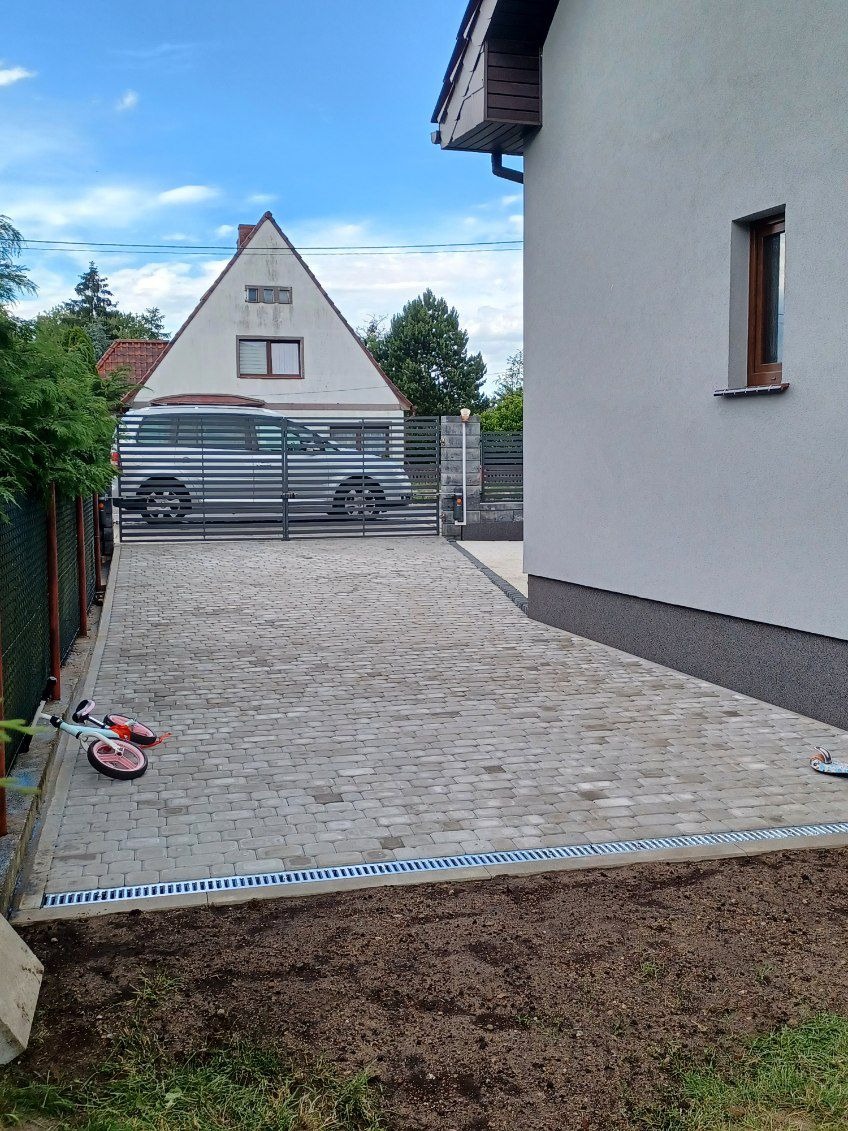 Ułożona szara kostka brukowa na podjeździe przed domem z metalową bramą, na pierwszym planie leży hulajnoga.