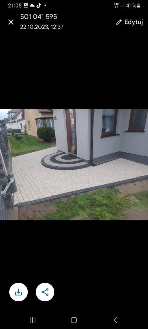 Ułożona jasna kostka brukowa prowadząca do wejścia domu z dekoracyjnym okręgiem z ciemnej kostki. Widoczne niedociągnięcia w postaci niezakończonych prac ziemnych przy obrzeżach.