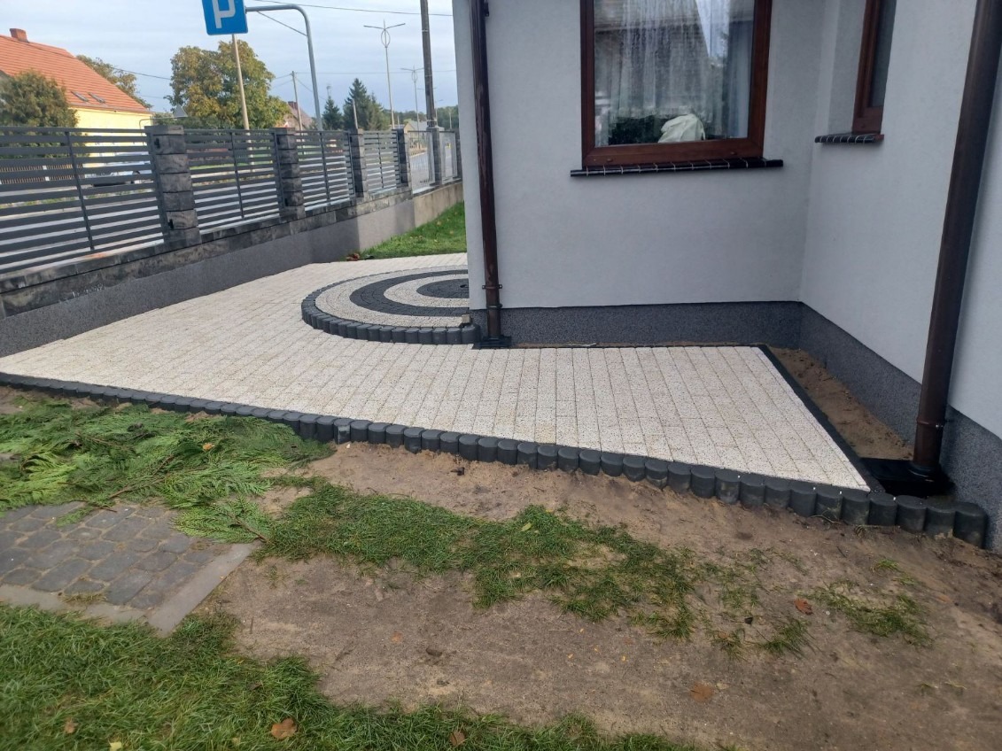 Ułożona kostka brukowa w kolorze beżowym z czarnymi obrzeżami, tworząca wzór koła przy ścianie budynku, widoczny fragment ogrodzenia panelowego i trawnik.