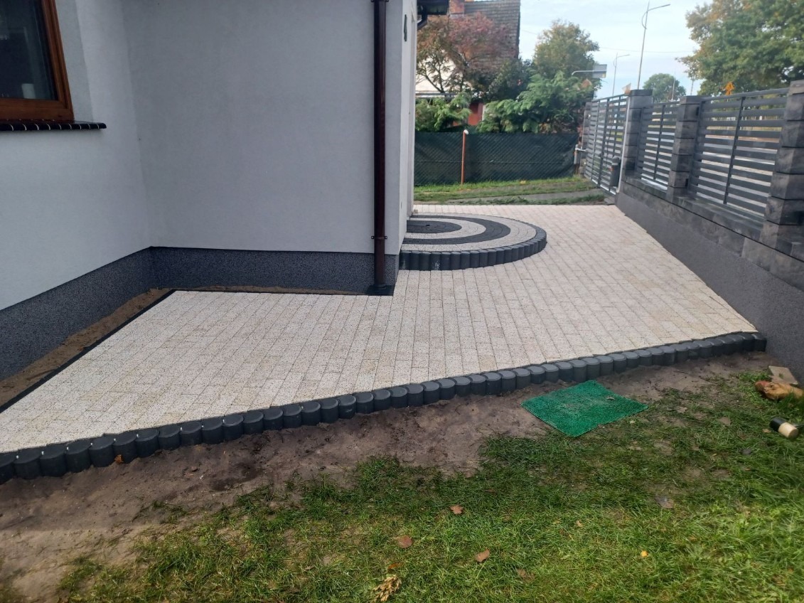 Ułożona jasna kostka brukowa z ciemnymi obrzeżami przy ścianie budynku i ogrodzeniu panelowym, z dekoracyjnym, półokrągłym wzorem z ciemnej kostki.