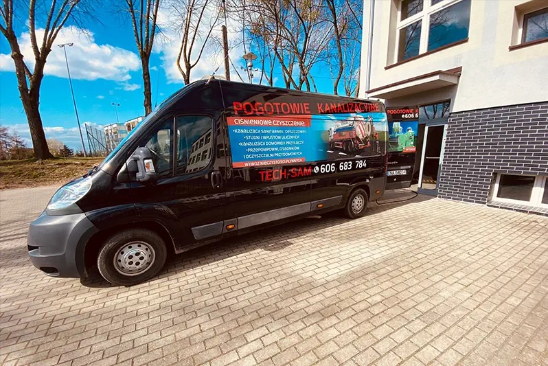 Czarny samochód dostawczy z napisem 'Pogotowie Kanalizacyjne' zaparkowany przed budynkiem z ciemnej cegły. Na samochodzie widoczne informacje o ciśnieniowym czyszczeniu kanalizacji, studni...