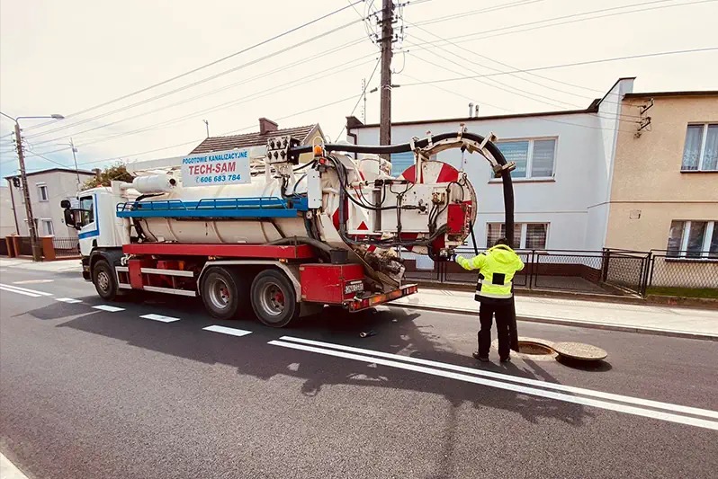 Pojazd asenizacyjny TECH-SAM z wężem skierowanym do studzienki kanalizacyjnej na ulicy, pracownik w żółtej kamizelce nadzoruje proces.