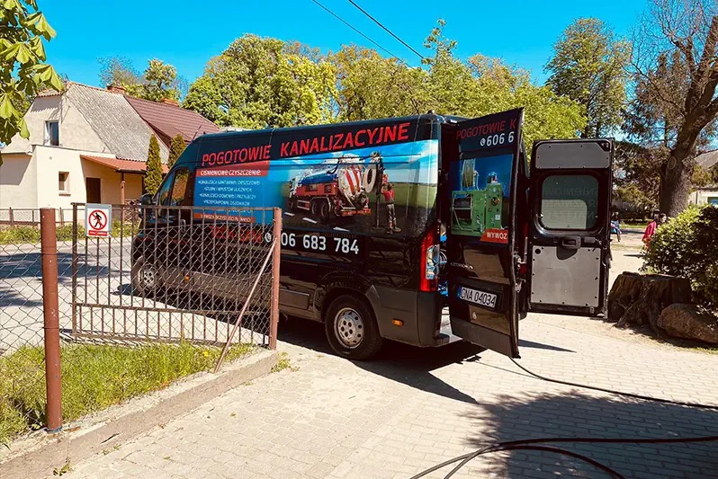 Czarny samochód dostawczy z otwartymi drzwiami, oklejony reklamą pogotowia kanalizacyjnego z numerem telefonu, zaparkowany na chodniku przed domem.
