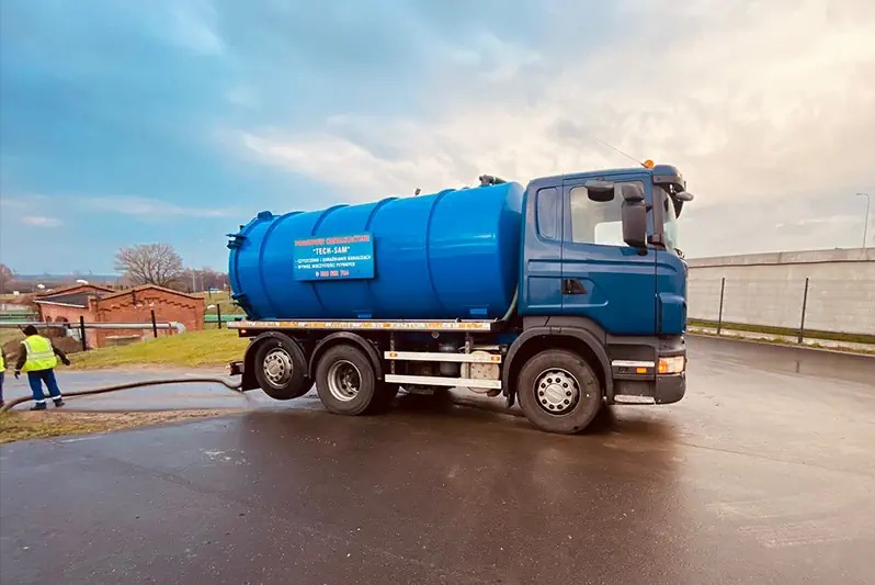 Niebieski samochód asenizacyjny 'TECH-SAM' z podłączonym wężem ssawnym, pracownik w żółtej kamizelce przy studzience kanalizacyjnej w tle ceglany budynek.