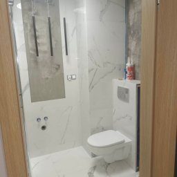 Mateusz Domański - Biała łazienka z marmurowymi płytkami, wisząca toaleta i minimalistyczne lampy wiszące, fragment ściany przygotowany pod płytki, widoczne pojemniki z silikonem.