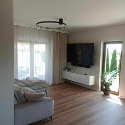 Mateusz Domański - Jasny salon z drewnianą podłogą, szarą sofą z poduszką, telewizorem na ścianie, białymi zasłonami i rośliną w doniczce przy drzwiach balkonowych.