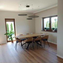 Mateusz Domański - Jasna jadalnia z drewnianym stołem, krzesłami z czarnymi nogami, okrągłymi lampami wiszącymi i podłogą z paneli, widok na zieleń przez okno.