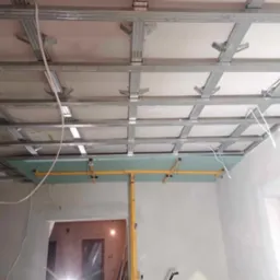 Montaż sufitu podwieszanego z płyt kartonowo-gipsowych, widoczne metalowe profile i fragment płyty podtrzymywanej przez żółty stempel budowlany.