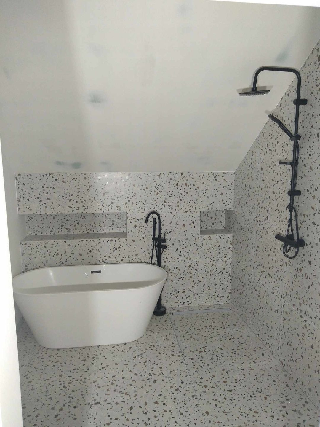 Nowoczesna łazienka w stylu lastryko z białą wanną wolnostojącą i czarną armaturą, wnęki w ścianie wykończone lastryko, sufit ze śladami wilgoci.