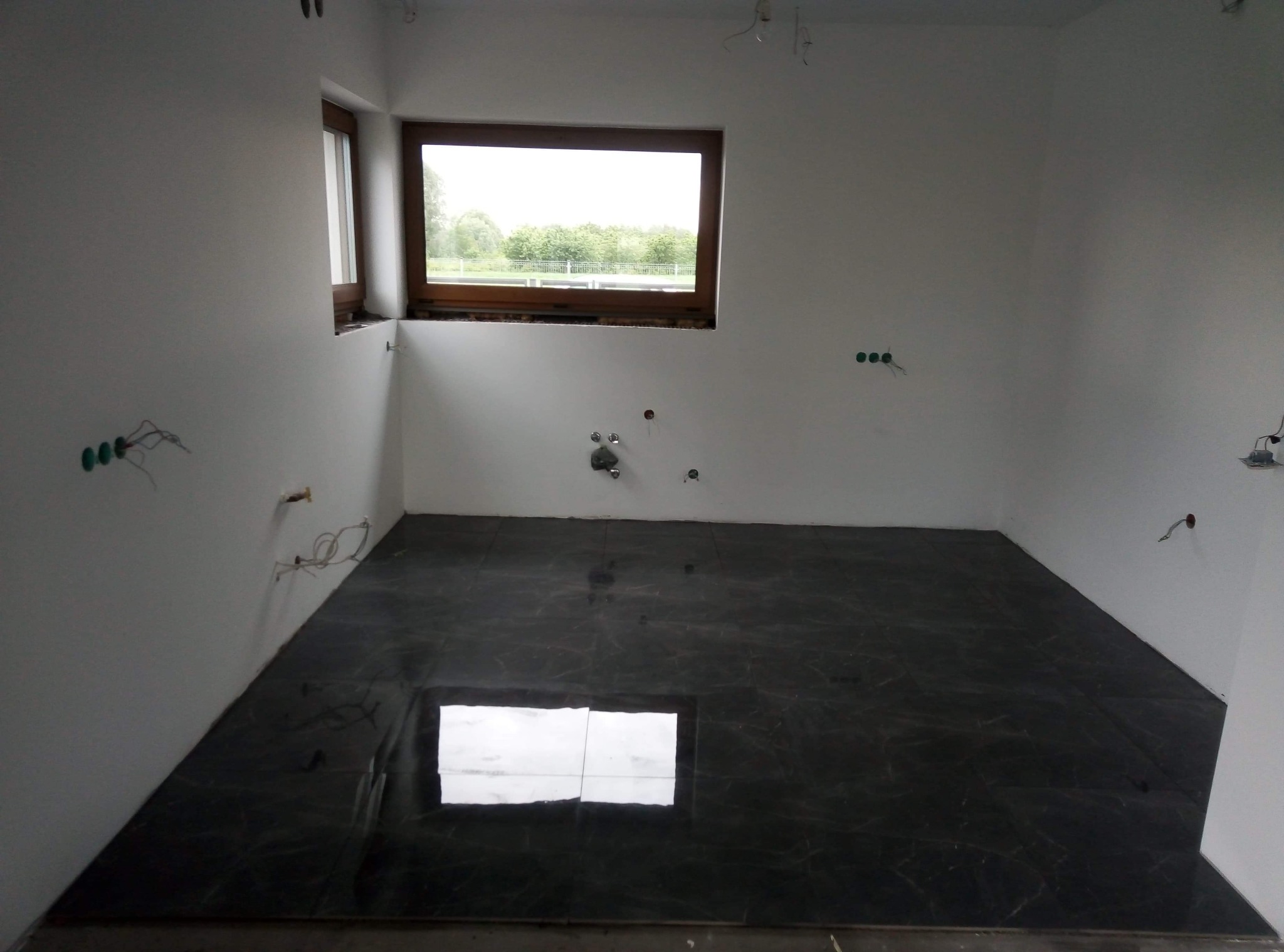 Łazienka w trakcie remontu: położona ciemna, marmurkowa podłoga z płytek, białe ściany z wystającymi kablami i rurami, okno z widokiem na zieleń.