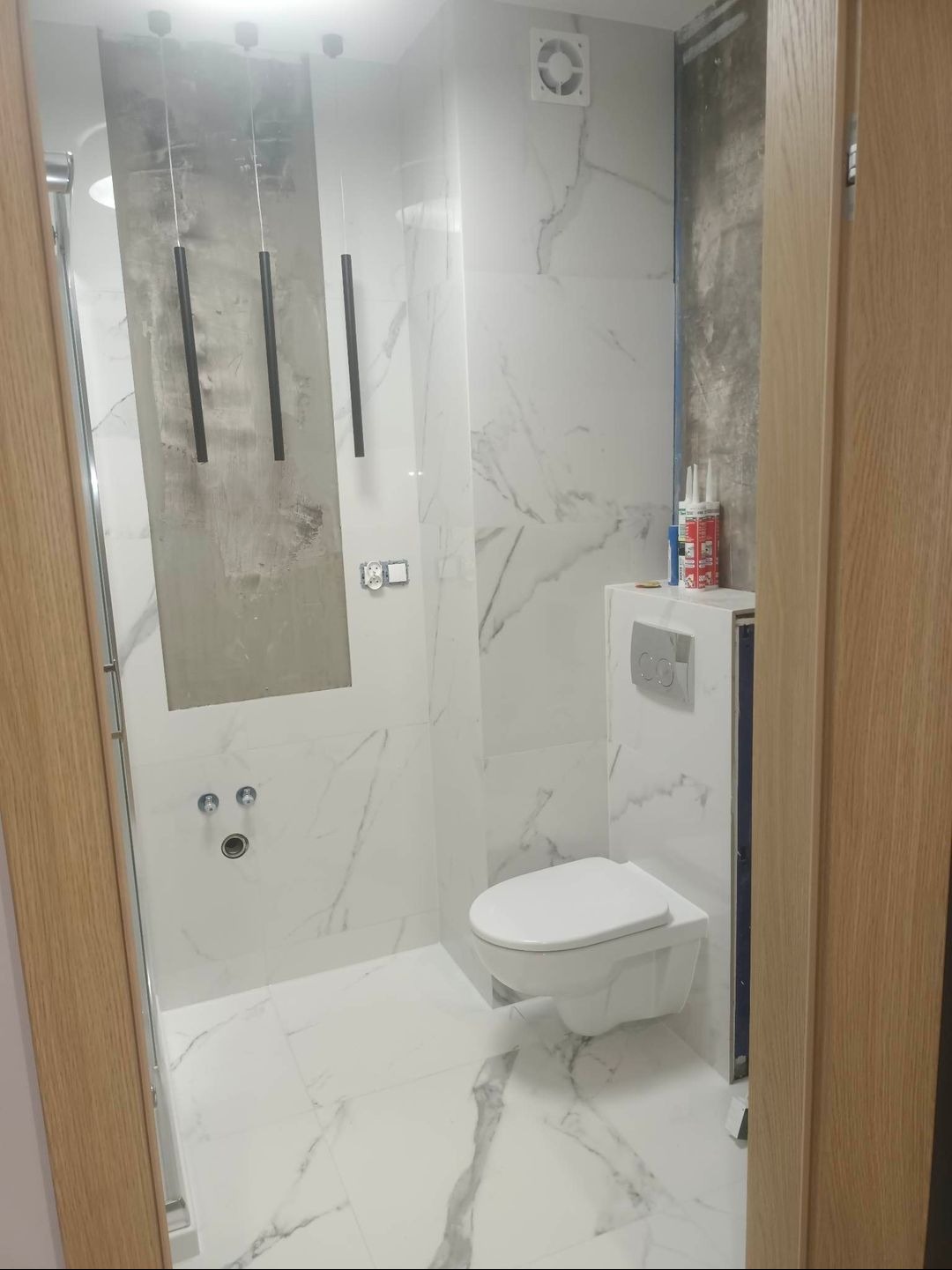 Biała łazienka z marmurowymi płytkami, wisząca toaleta i minimalistyczne lampy wiszące, fragment ściany przygotowany pod płytki, widoczne pojemniki z silikonem.