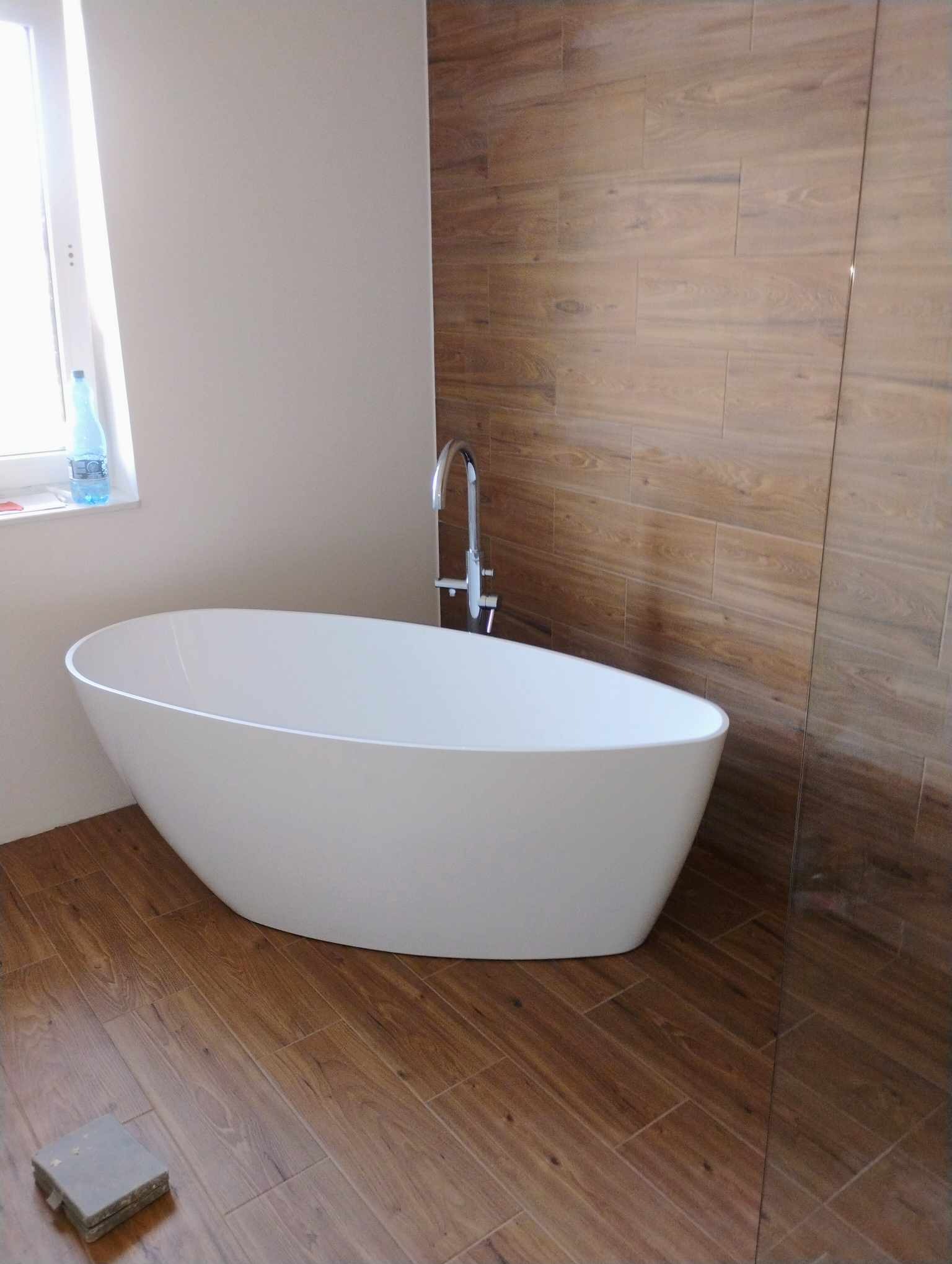 Wolnostojąca, owalna wanna z chromowaną baterią, umieszczona w wykończonej łazience z drewnopodobnymi płytkami na ścianach i podłodze. W tle okno z butelką wody mineralnej.