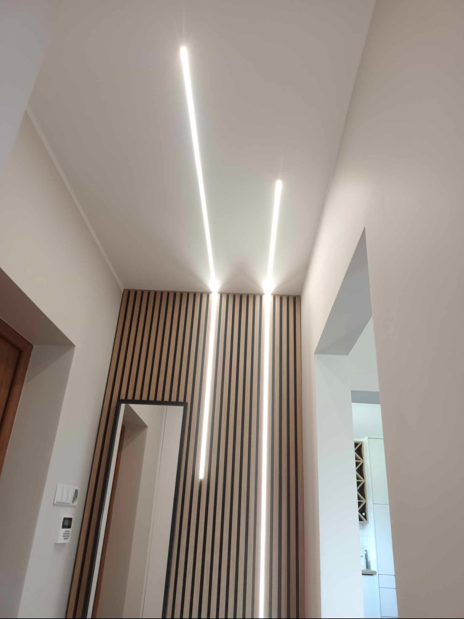 Nowoczesny korytarz z drewnianą ścianą z pionowych listew, oświetleniem LED wpuszczonym w sufit i dużym lustrem stojącym.