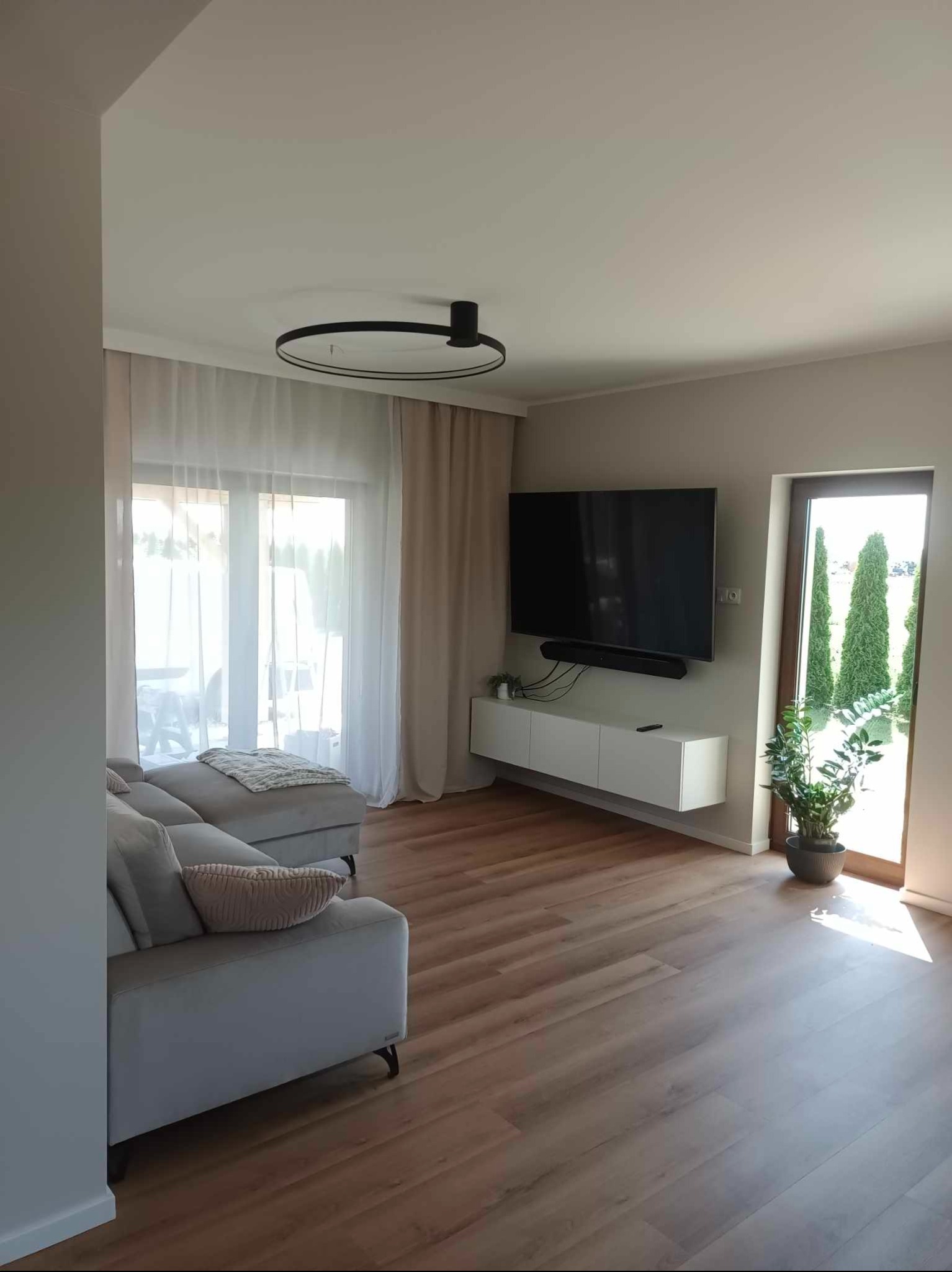 Jasny salon z drewnianą podłogą, szarą sofą z poduszką, telewizorem na ścianie, białymi zasłonami i rośliną w doniczce przy drzwiach balkonowych.