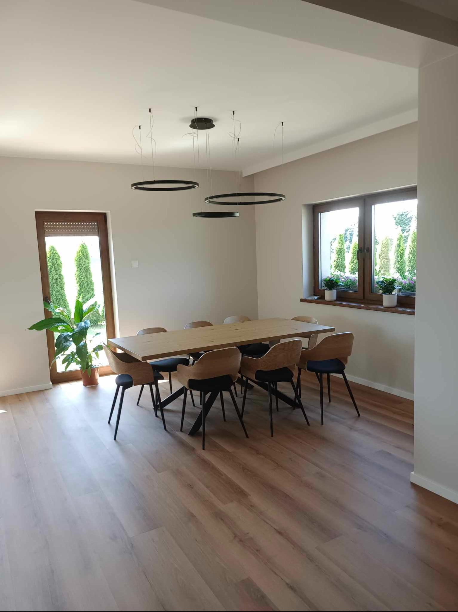 Jasna jadalnia z drewnianym stołem, krzesłami z czarnymi nogami, okrągłymi lampami wiszącymi i podłogą z paneli, widok na zieleń przez okno.