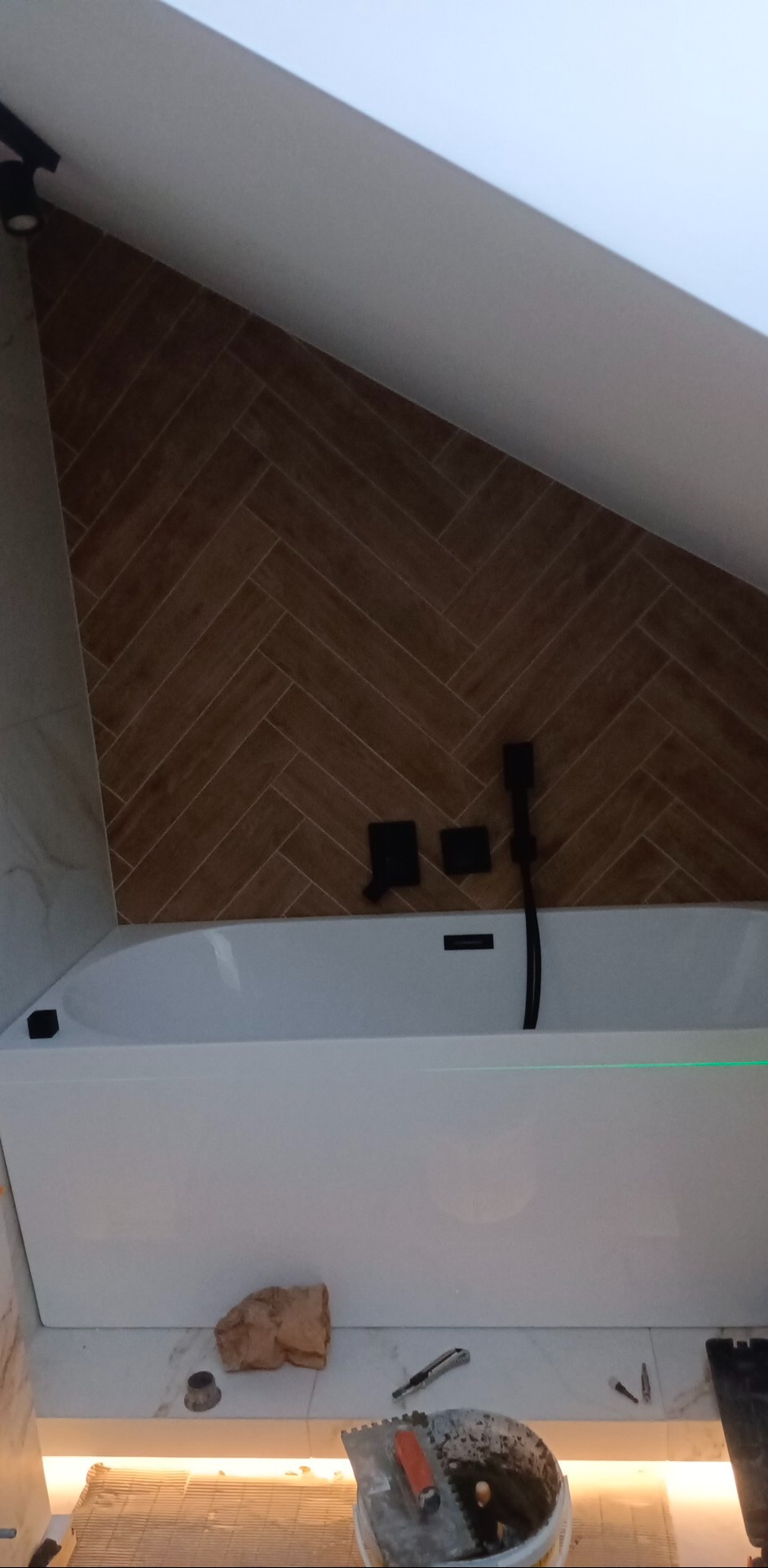 Ukośna ściana łazienki z brązowymi płytkami ułożonymi w jodełkę nad białą wanną, czarna armatura i poziomica laserowa, widoczne narzędzia glazurnicze.