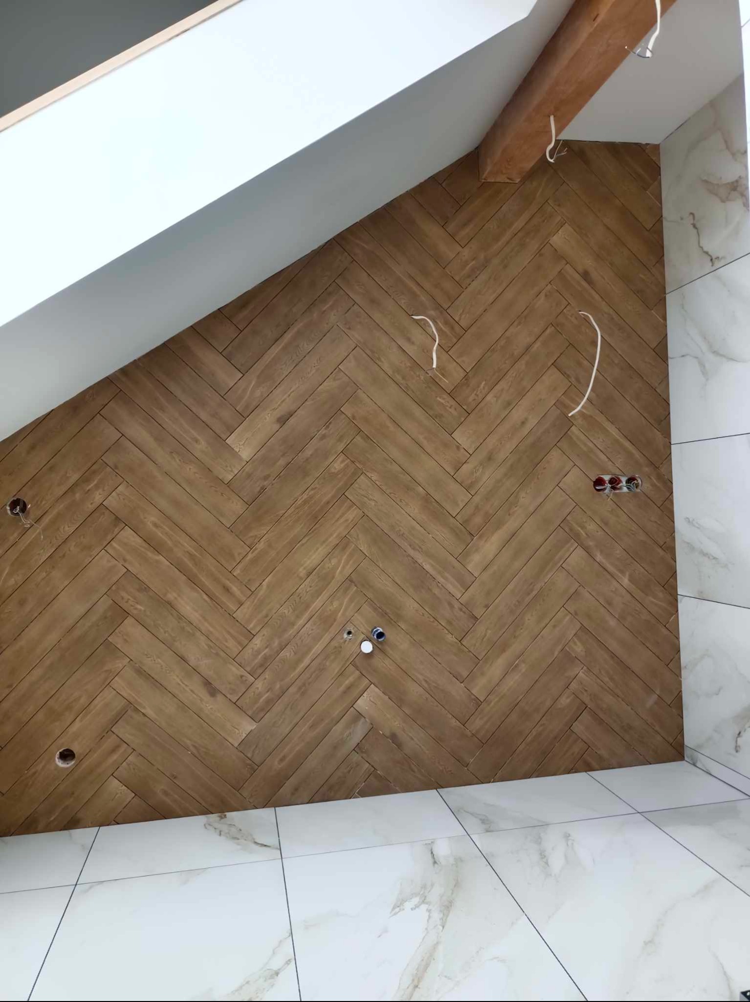 Ściana wykończona drewnopodobną okładziną w jodełkę, widoczne wystające przewody elektryczne i otwory na gniazdka, białe marmurowe płytki na podłodze i częściowo na ścianie, skośny sufit...