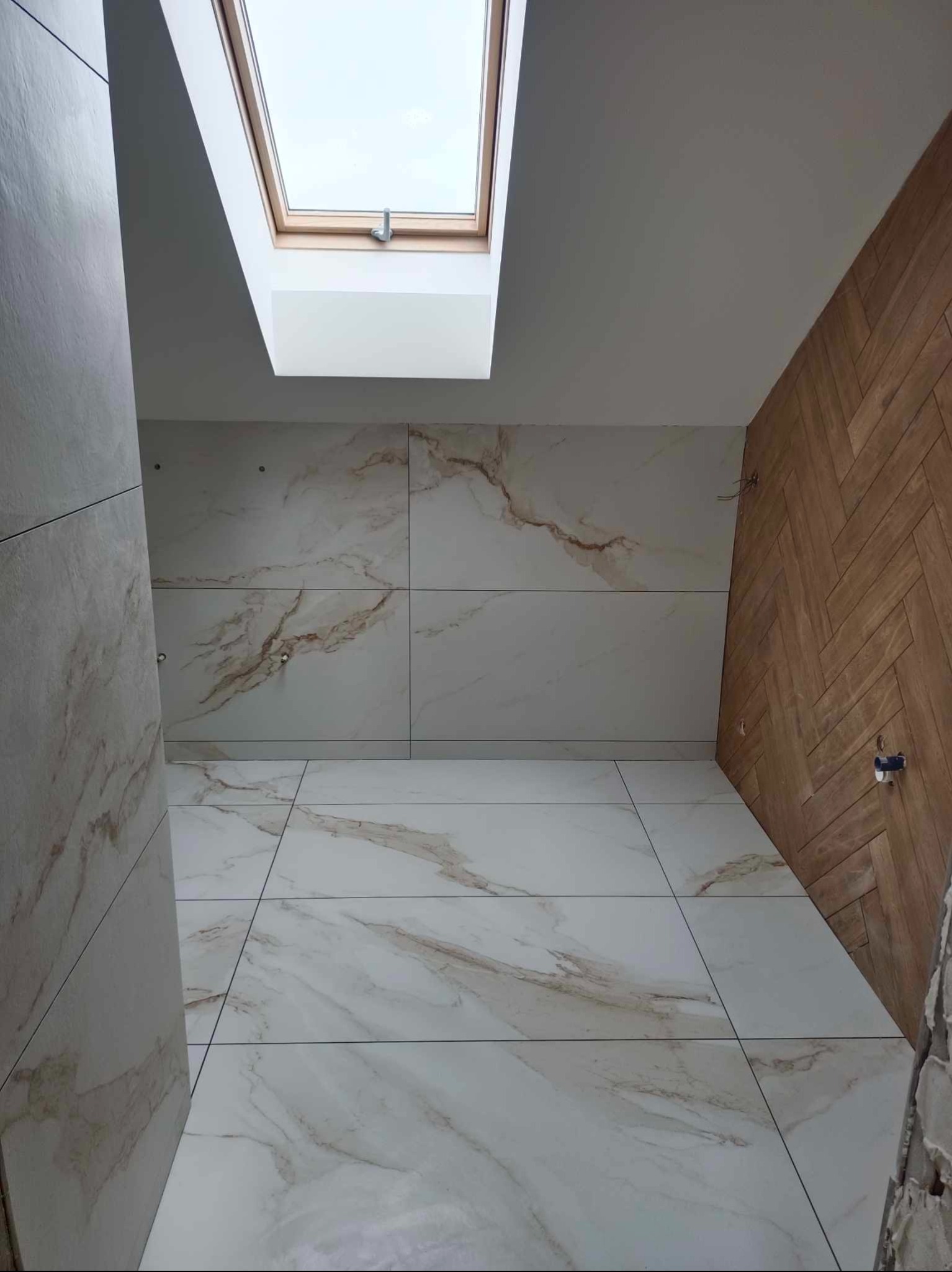 Wykończona łazienka z marmurowymi płytkami na podłodze i ścianach, drewnianą ścianą ułożoną w jodełkę oraz oknem dachowym wpuszczającym naturalne światło.