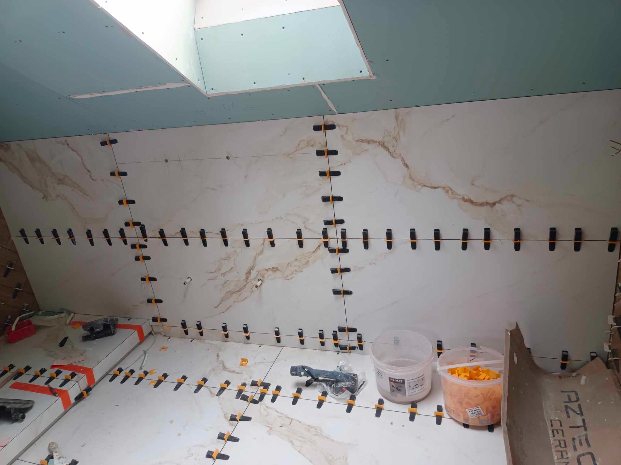 Ściana i podłoga w trakcie układania płytek imitujących marmur, z użyciem systemu poziomowania płytek, widoczne narzędzia i materiały, sufit z płyt kartonowo-gipsowych.