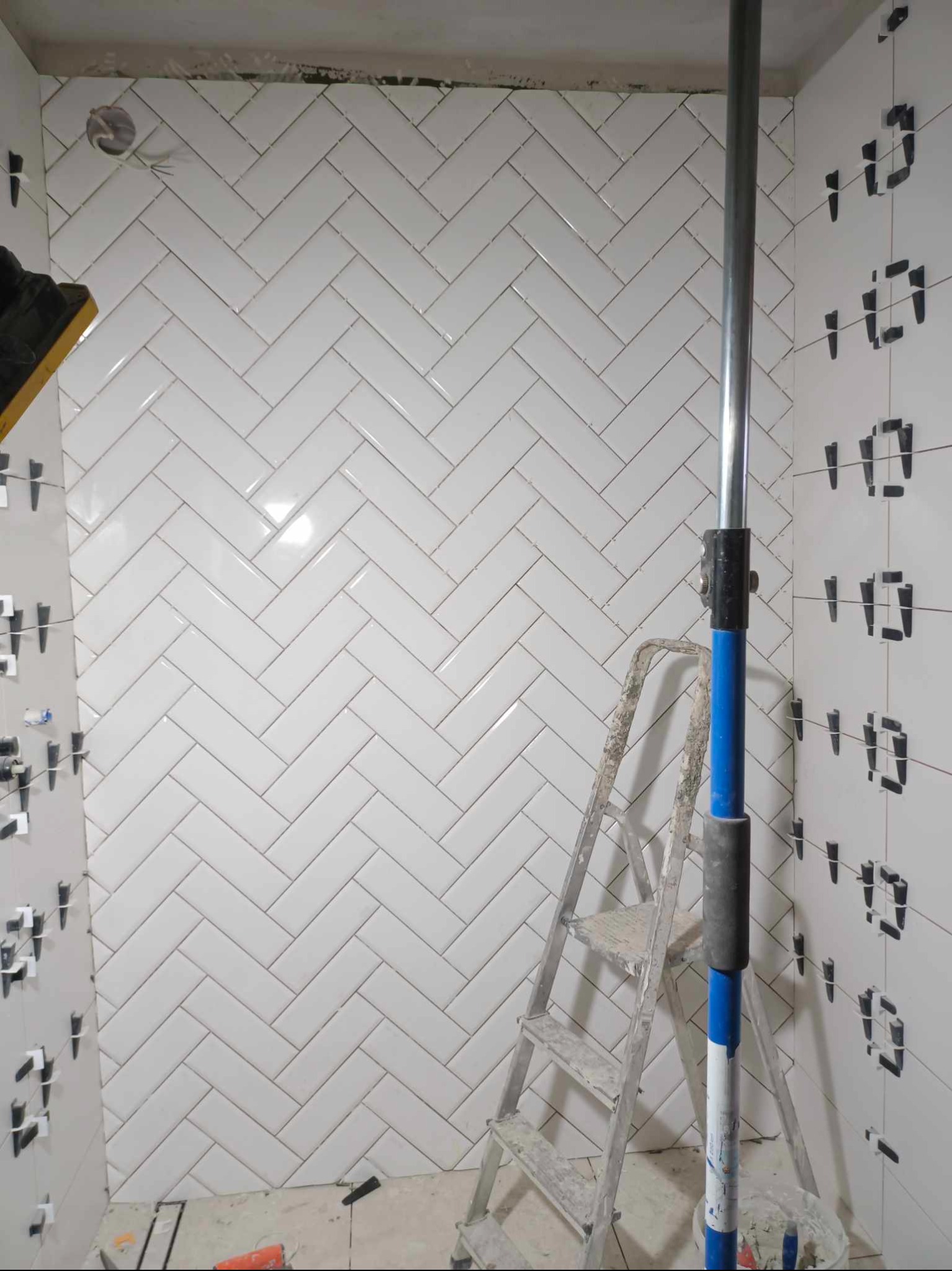 Ściana łazienki w trakcie układania białych, błyszczących płytek w jodełkę, widoczne krzyżyki dystansowe, drabina malarska i rozpórka sufitowa.