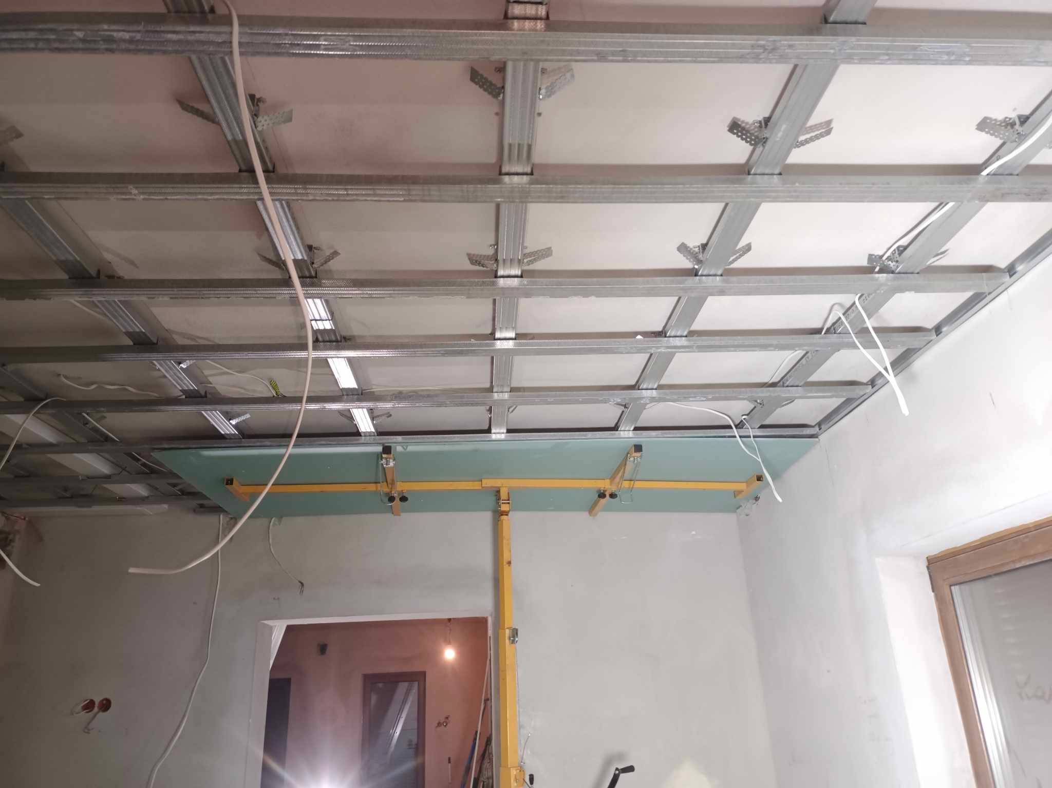 Montaż sufitu podwieszanego z płyt kartonowo-gipsowych, widoczne metalowe profile i fragment płyty podtrzymywanej przez żółty stempel budowlany.