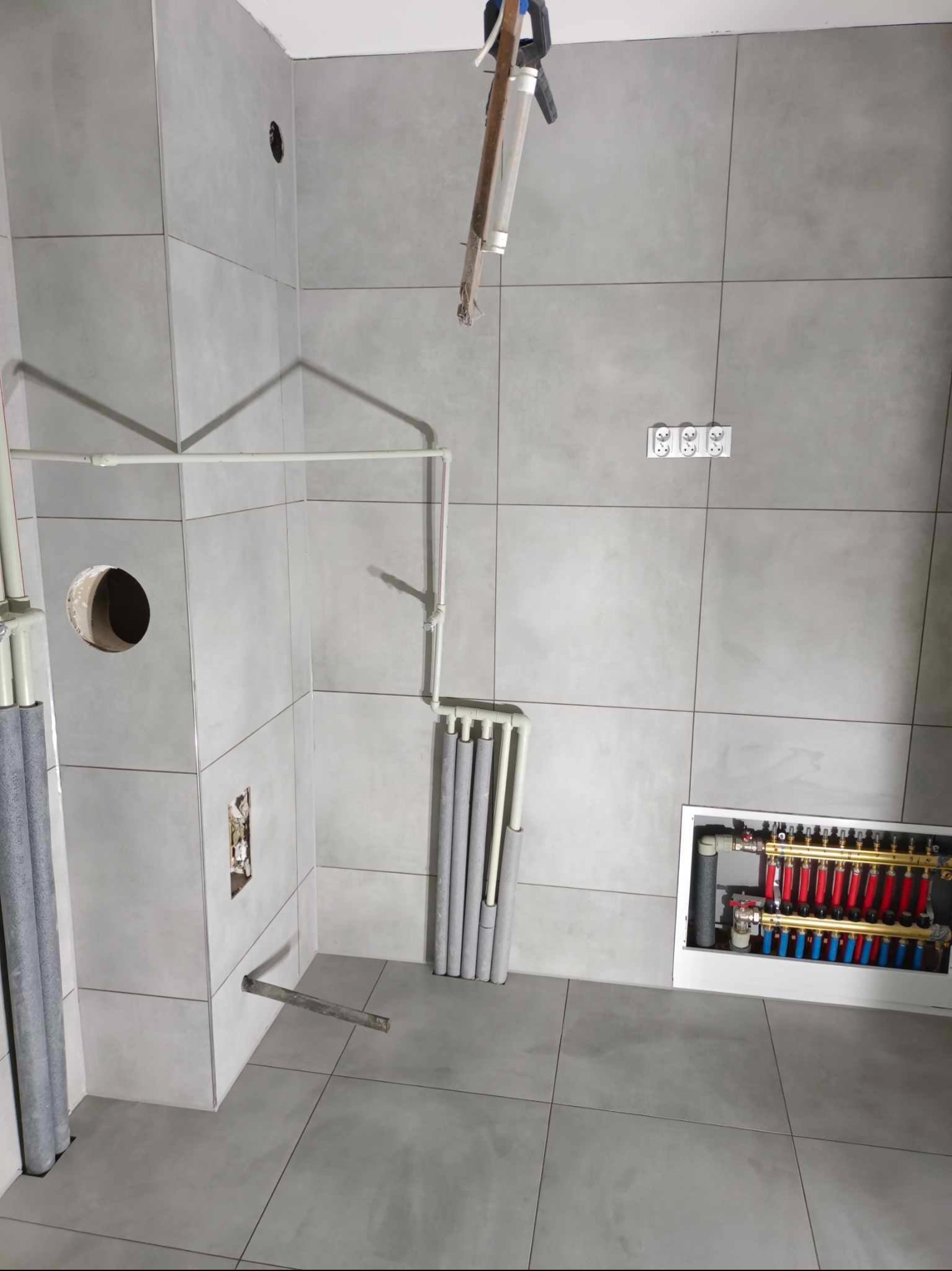 Remontowana łazienka w Sieradzu: szare płytki, białe rury, rozdzielacz ogrzewania podłogowego w zabudowie, widoczne gniazdka elektryczne i otwór w ścianie.