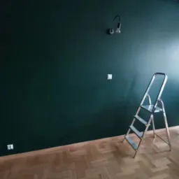 Świeżo pomalowana ściana w odcieniu butelkowej zieleni z widoczną drabiną aluminiową, gniazdkiem elektrycznym i lampą ścienną w pomieszczeniu z parkietem.