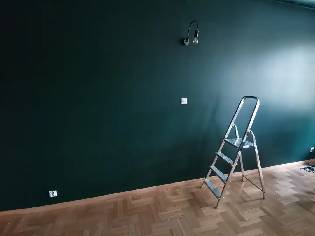 Świeżo pomalowana ściana w odcieniu butelkowej zieleni z widoczną drabiną aluminiową, gniazdkiem elektrycznym i lampą ścienną w pomieszczeniu z parkietem.