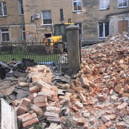 ROZBIÓRKI WYBURZENIA - Ceglany gruz po rozbiórce na tle budynku z odsłoniętą cegłą i żółtą koparką za ogrodzeniem. Widok z bliska na plac budowy.