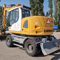 ROZBIÓRKI WYBURZENIA - Żółto-biała koparka Liebherr 914 Compact na kołach, z łyżką z przodu i lemieszem z tyłu, na tle drzew i nieba. Widoczne detale: opony, kabina, oznaczenia.