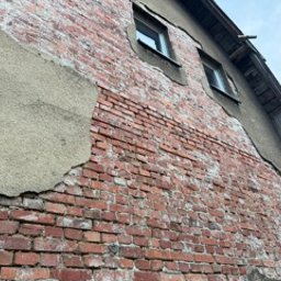 ROZBIÓRKI WYBURZENIA - Odsłonięta ceglana ściana budynku z fragmentami tynku, widoczne okna i drewniana konstrukcja dachu. Ujęcie z dołu, podkreślające strukturę i fakturę materiałów.