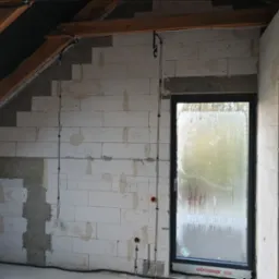 Surowy stan poddasza z widocznymi przewodami elektrycznymi w ścianach z betonu komórkowego, okno z zaparowaną szybą i drewniane elementy konstrukcyjne dachu.