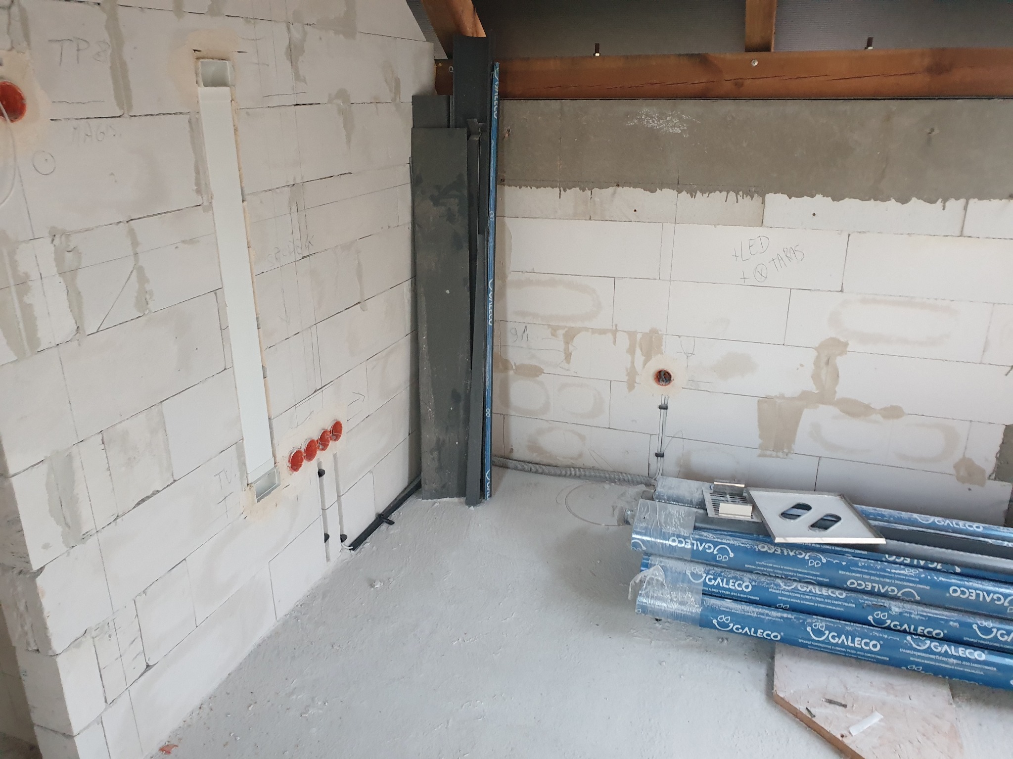 Surowy stan wnętrza w trakcie prac: widoczne instalacje elektryczne w ścianach z betonu komórkowego, rury osłonowe Galeco, puszki elektryczne w kolorze czerwonym i przewody. Ściany częściowo...