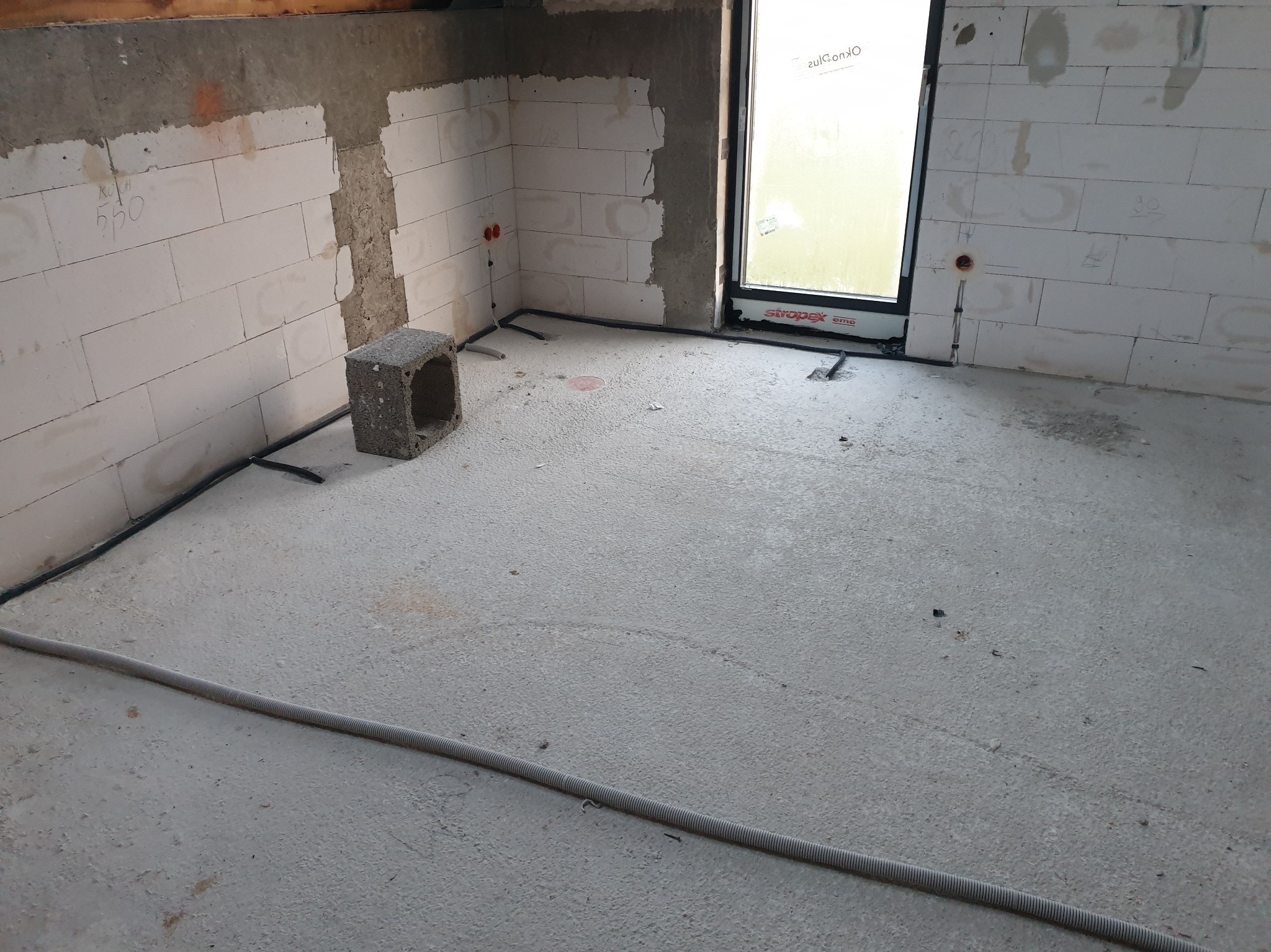 Wnętrze w stanie surowym z widocznymi przewodami elektrycznymi ułożonymi na podłodze i wypustami na ścianach z bloków betonowych, przygotowane do dalszych prac wykończeniowych.