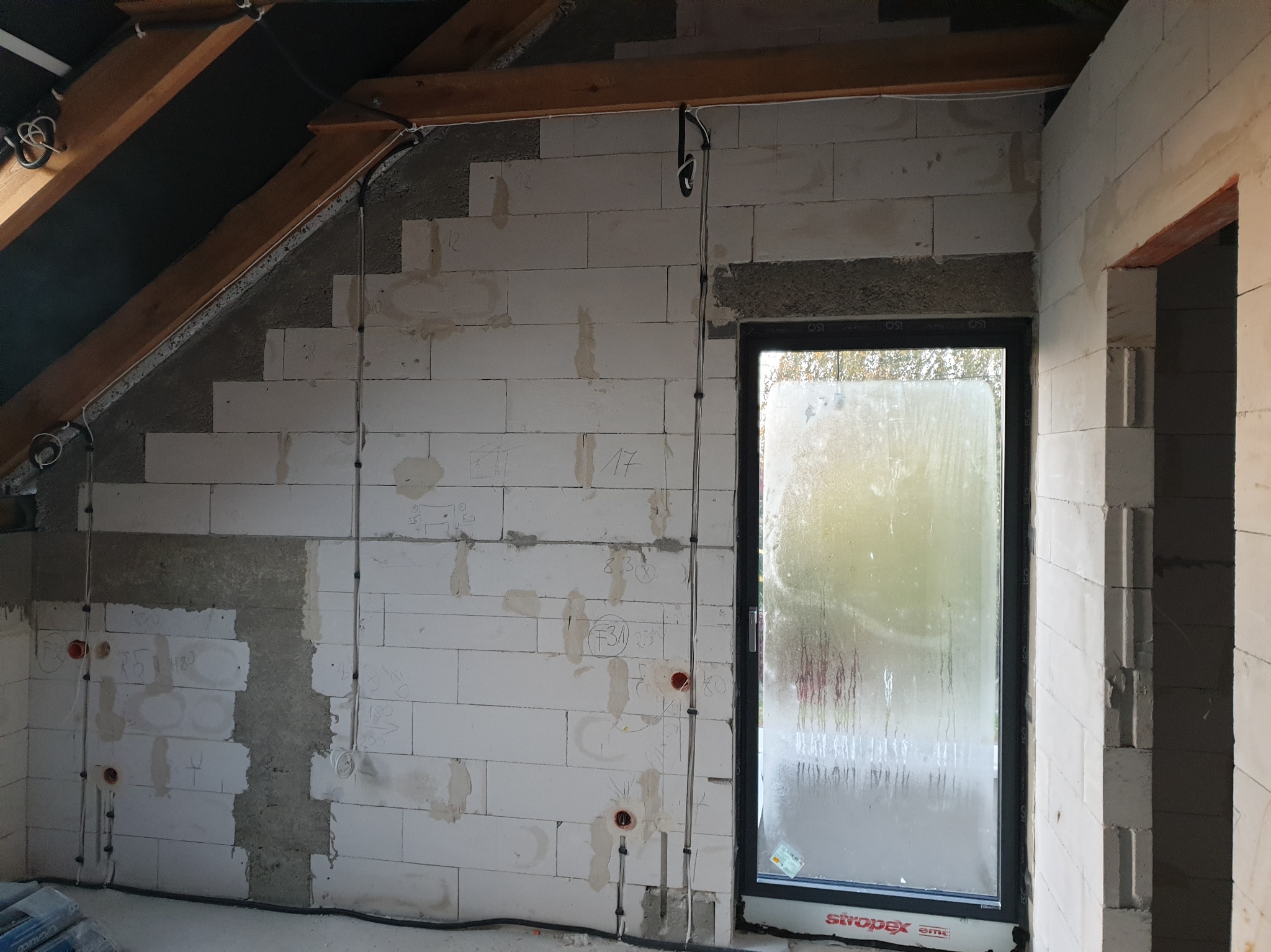 Wewnętrzna ściana z betonu komórkowego w trakcie prac elektrycznych: widoczne kable, puszki instalacyjne, bruzdy oraz oznaczenia na ścianie, okno z widocznymi kroplami wody.