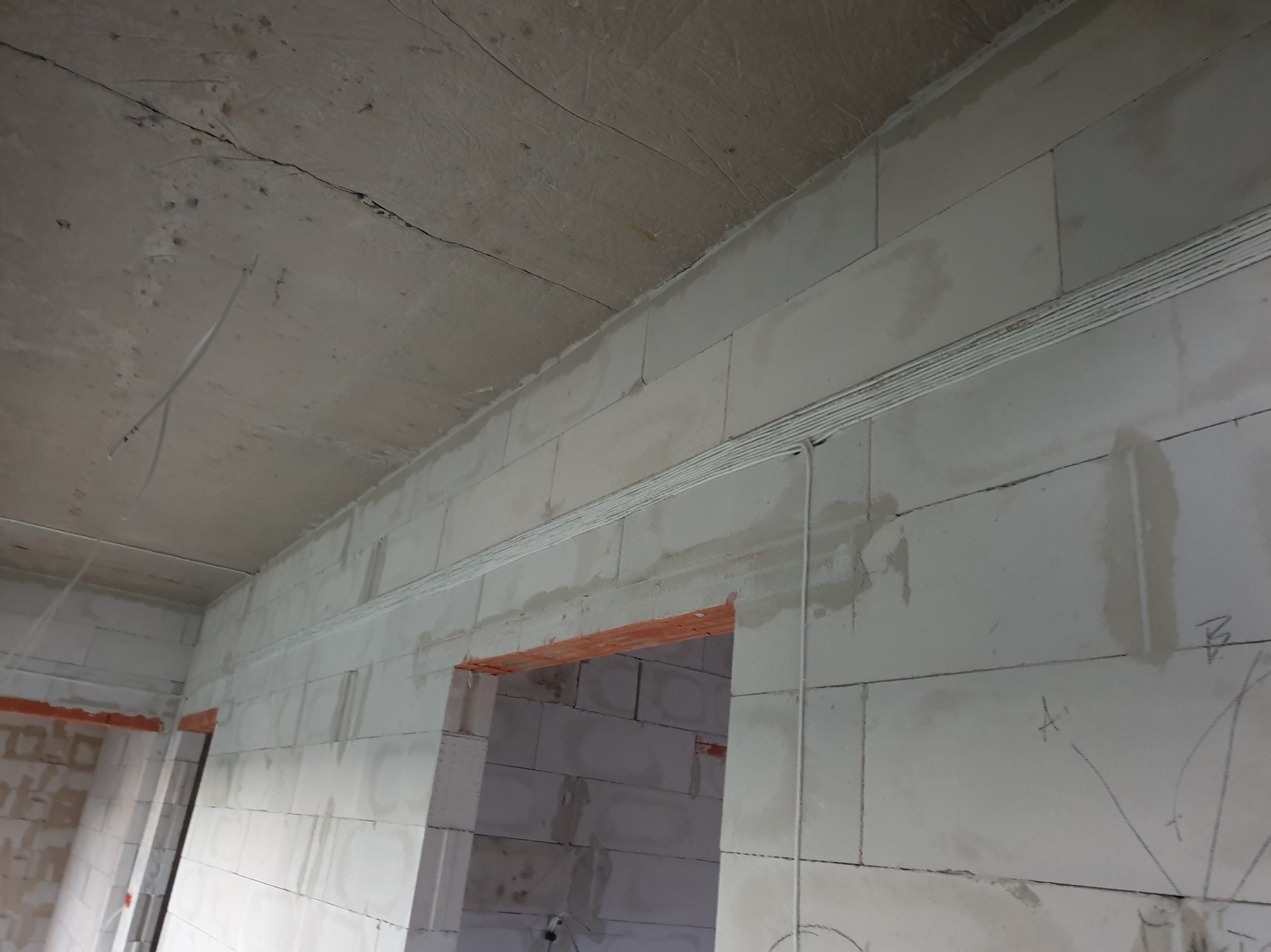 Wnętrze w trakcie budowy z widocznymi przewodami elektrycznymi ułożonymi w korycie nad otworem drzwiowym, ściany z białych bloczków, surowy betonowy strop.