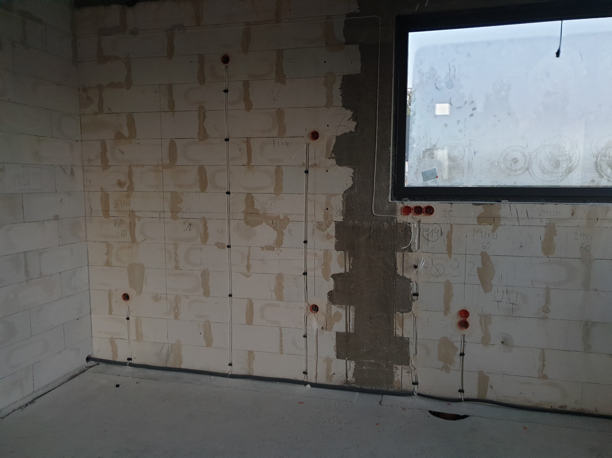 Surowy stan: instalacja elektryczna w ścianie z betonu komórkowego, widoczne przewody w peszlach i puszki podtynkowe, okno z brudną szybą.