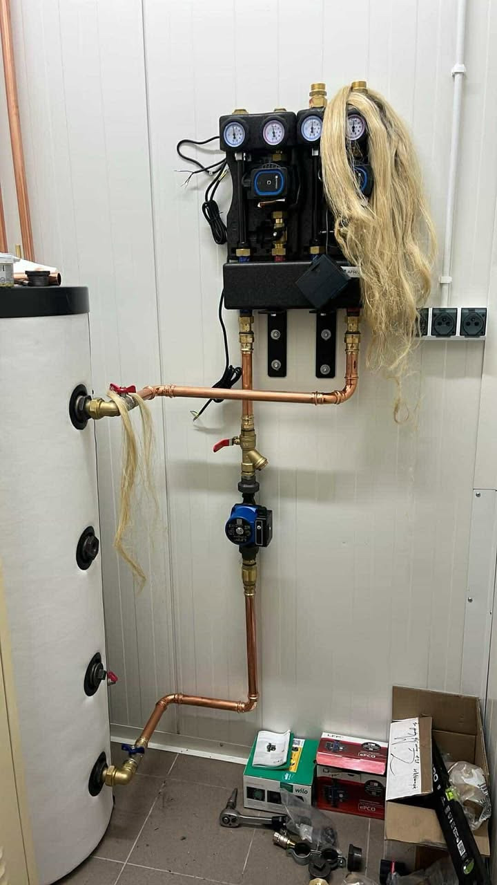 Instalacja CO z kotłem i osprzętem, rury miedziane, manometry. Na instalacji i kotle leżą blond włosy. Na podłodze narzędzia i pudełka po osprzęcie.