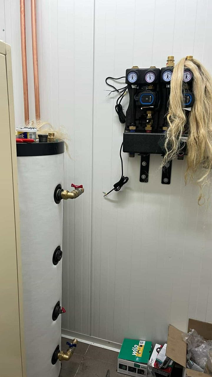 Bojler z podłączoną armaturą i rozdzielaczem CO na ścianie w pomieszczeniu technicznym. Na rozdzielaczu leży blond peruka. Widoczne narzędzia i akcesoria.