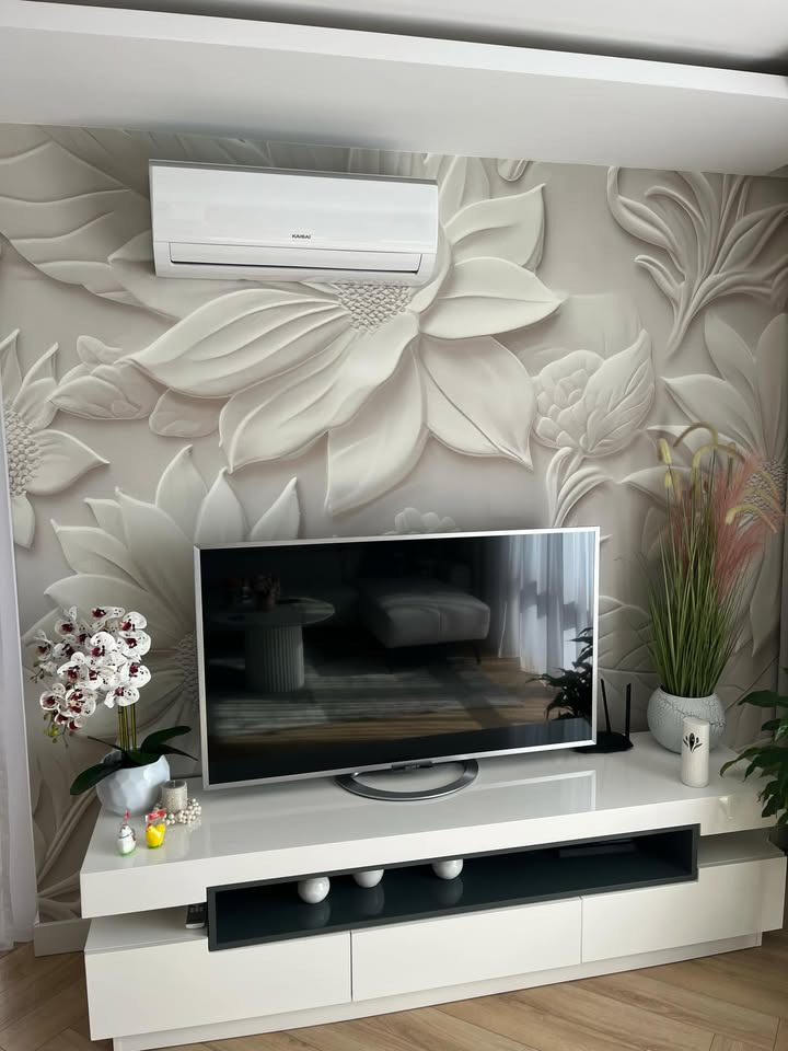Biały klimatyzator marki Kaisai na ścianie z kwiatowym wzorem 3D nad telewizorem i szafką RTV w nowoczesnym salonie. Widoczne dekoracje i rośliny.