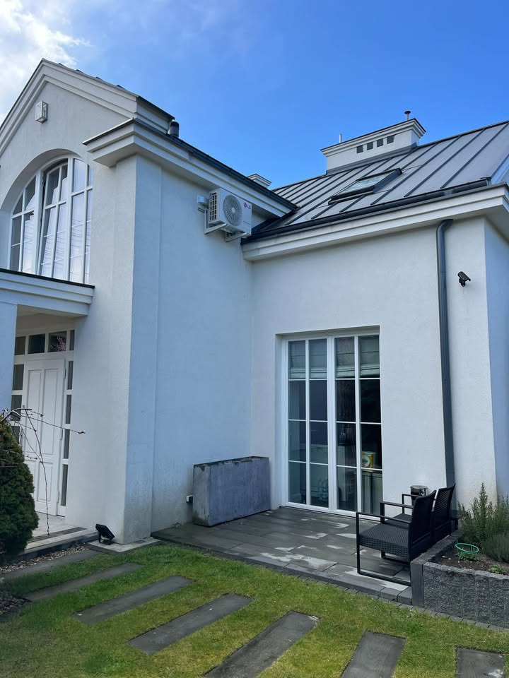 Biały dom z szarym dachem, widoczne okna, drzwi, trawnik z betonowymi płytami, klimatyzator na ścianie i kamera. Elegancki minimalizm.