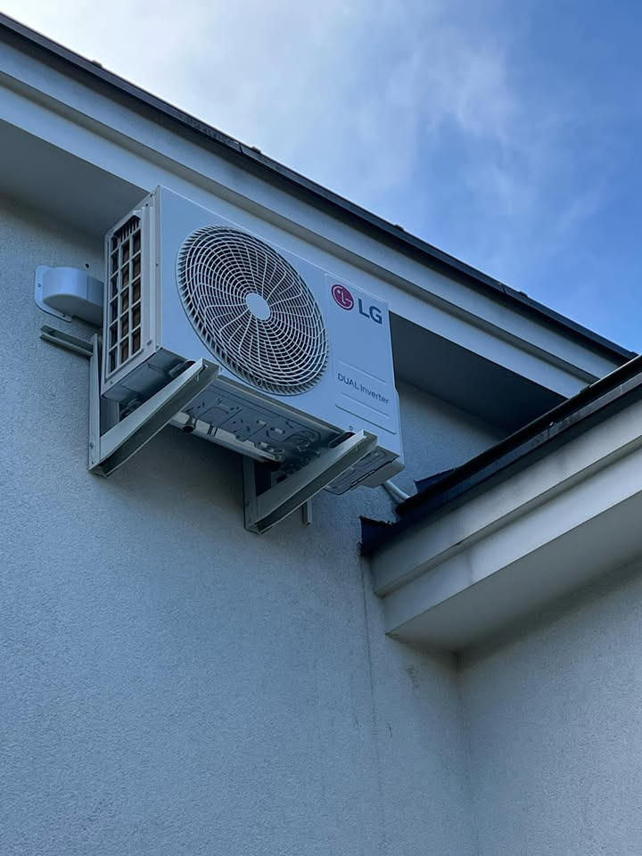 Biała jednostka zewnętrzna klimatyzacji LG Dual Inverter zamontowana na ścianie budynku z widocznymi wspornikami i fragmentem dachu.