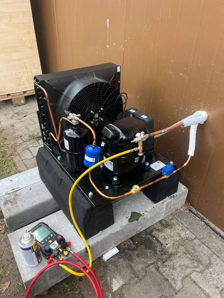 Czarna sprężarka chłodnicza na betonowym podłożu, podłączona do instalacji, z widocznymi narzędziami pomiarowymi i pianką uszczelniającą na rurze przy ścianie.