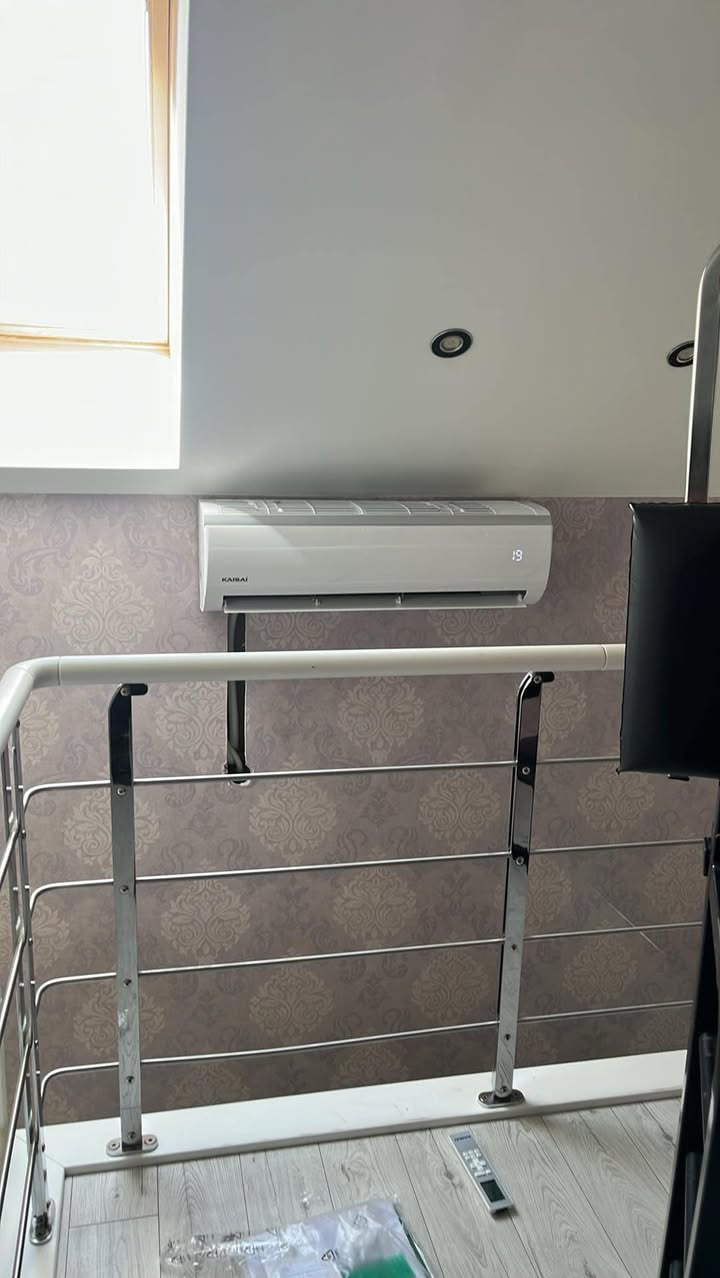 Nowoczesny klimatyzator marki Kaisai zamontowany na ścianie z wzorzystą tapetą, widoczny zza metalowej barierki. Na podłodze pilot i folia.