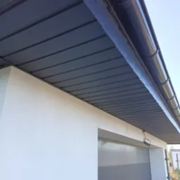 Szara podbitka dachowa z blachy trapezowej i rynna w kolorze grafitowym zamontowane przy białej elewacji budynku.