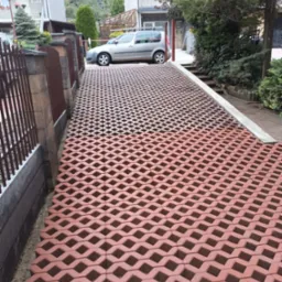 Ułożona, ażurowa nawierzchnia podjazdu z czerwonej kostki na tle częściowo widocznego ogrodzenia, domu i zaparkowanego samochodu.