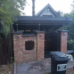 Zadaszone miejsce na pojemniki na śmieci z ceglanymi ściankami, ozdobnym okienkiem i metalową bramką, obok drewniany płot i pojemnik na odpady komunalne.