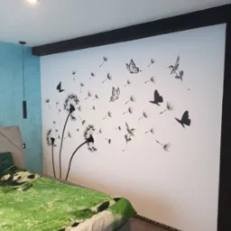 Biała ściana w sypialni z naklejonym wzorem dmuchawców, motyli i kolibrów w kolorze czarnym, widoczne oświetlenie sufitowe i fragment łóżka z zieloną pościelą.