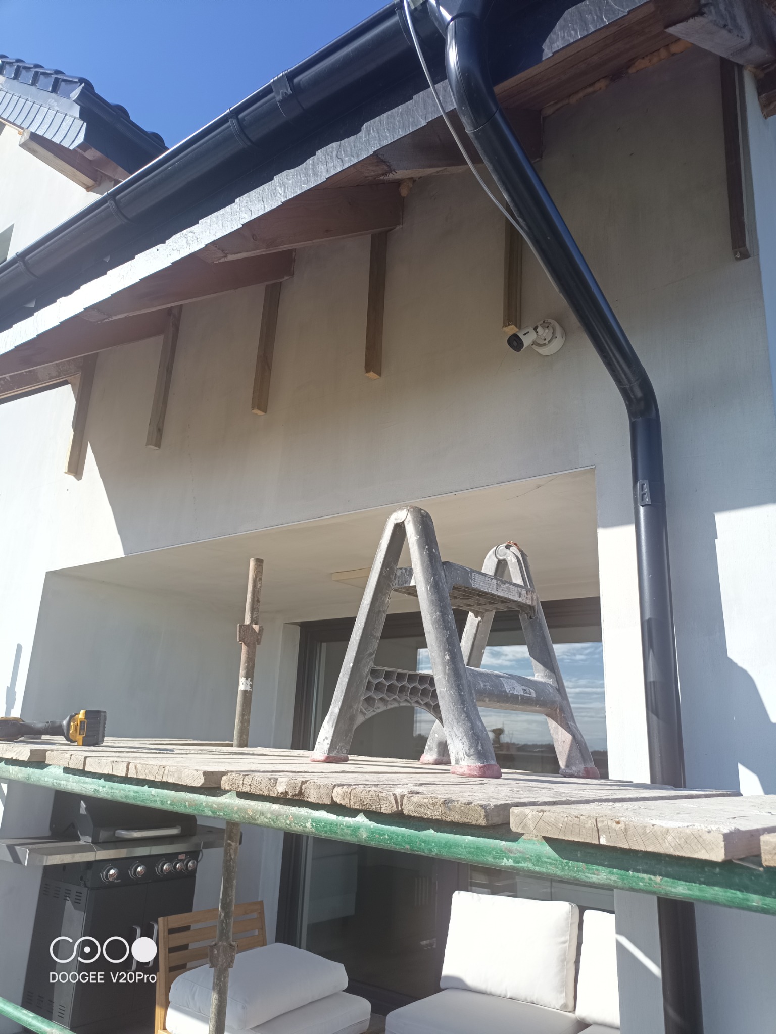 Elewacja budynku w trakcie prac wykończeniowych, widoczne rusztowanie, drabina, przygotowane deski, czarna rynna oraz kamera monitoringu.