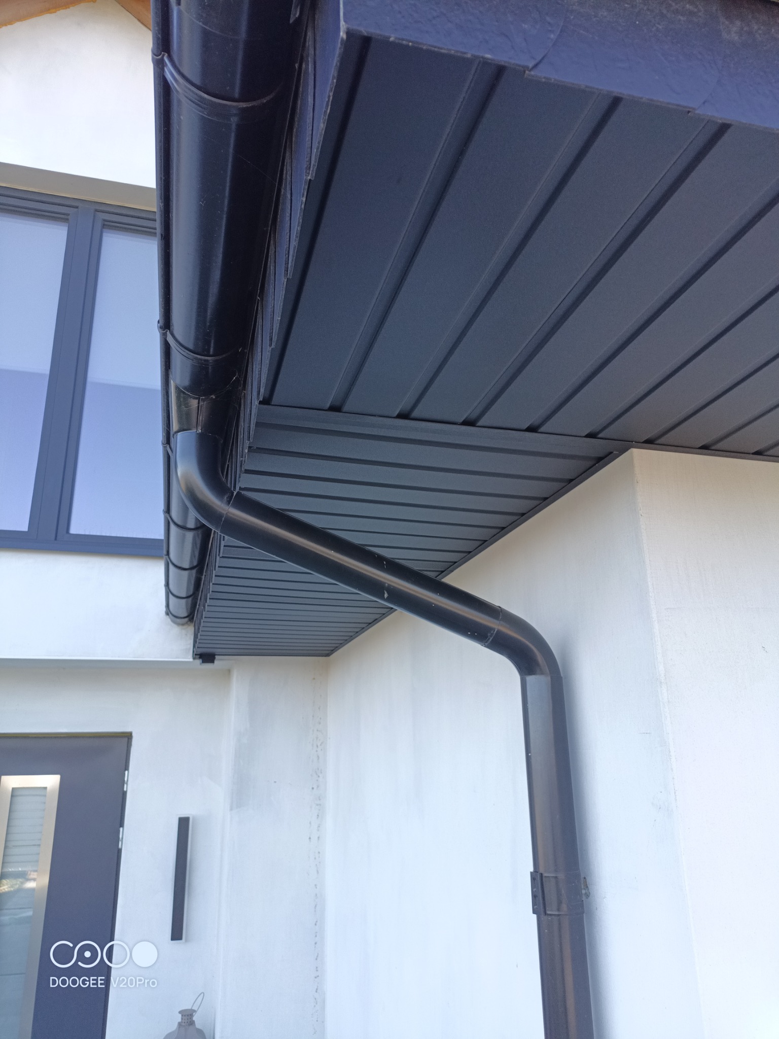 Ciemnoszara podbitka dachowa z orynnowaniem w kolorze, widok od dołu, fragment elewacji z drzwiami wejściowymi i oknem.